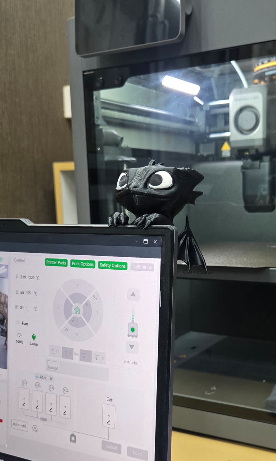 A printed dragon sitting on a laptop lid, the printer running behind it in the shop.